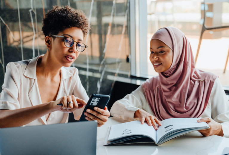 Zwei Frauen lernen gemeinsam mit Smartphone und Buch in moderner Umgebung für eine Sprachweiterbildung – TASys